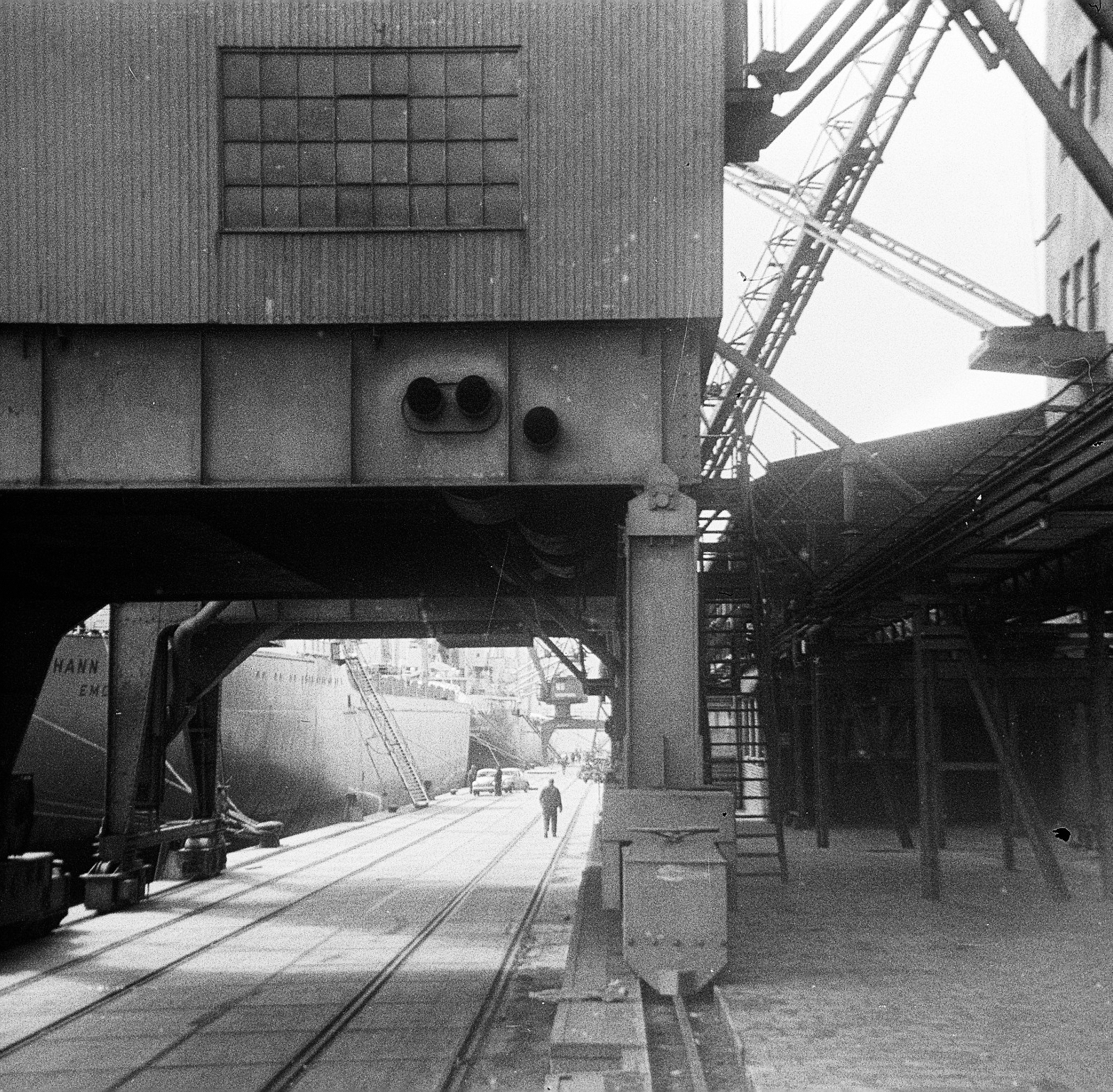 Blick unter einm großen Schiffskran hindurch auf VW-Käfer, die auf die MS Johann Schulte verladen und in die USA transportiert werden sollen. Die MS Johann Schulte mit Gangway. 1967. Foto: Erwin Thomasius.